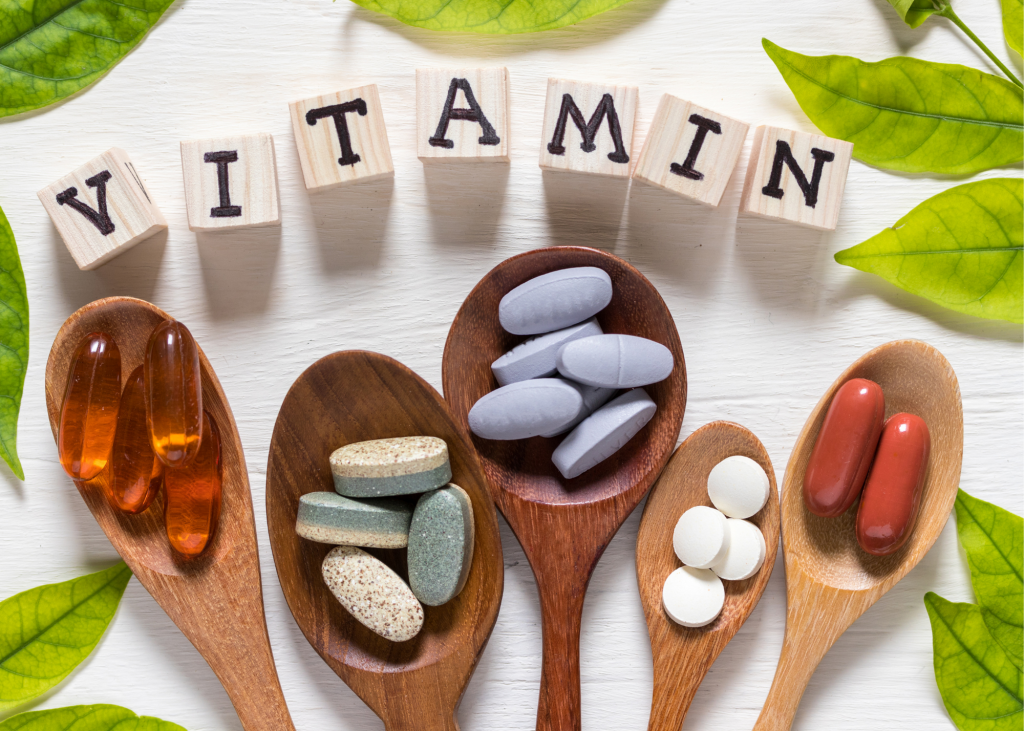 Five spoons with beneficial vitamins in them on a white table with blocks spelling out "vitamin," and green leaves surrounding both.