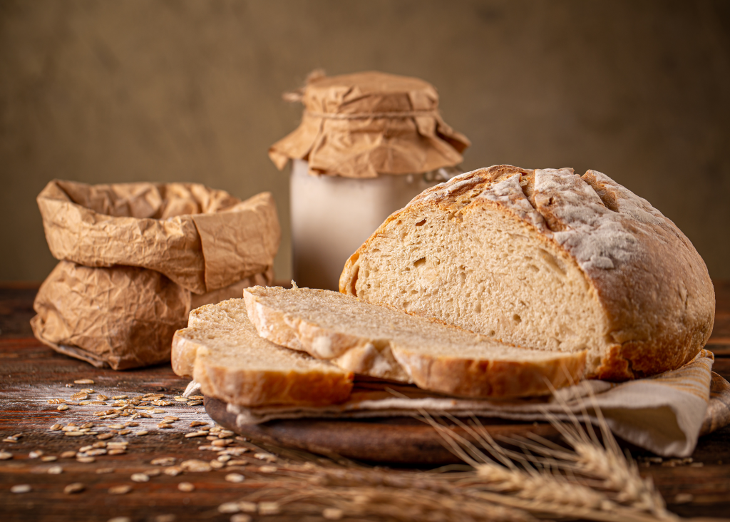 Fermented sourdough loaf partially sliced on cutting board. Sourdough started in jar behind it with a brown paper bag beside it. Grain scattered on brown table with whole grain stalks visible in front.