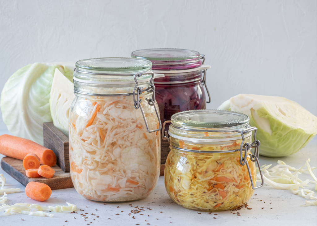 Three jars full of scientifically researched sauerkraut with cabbage, carrot and seeds behind and on the side.