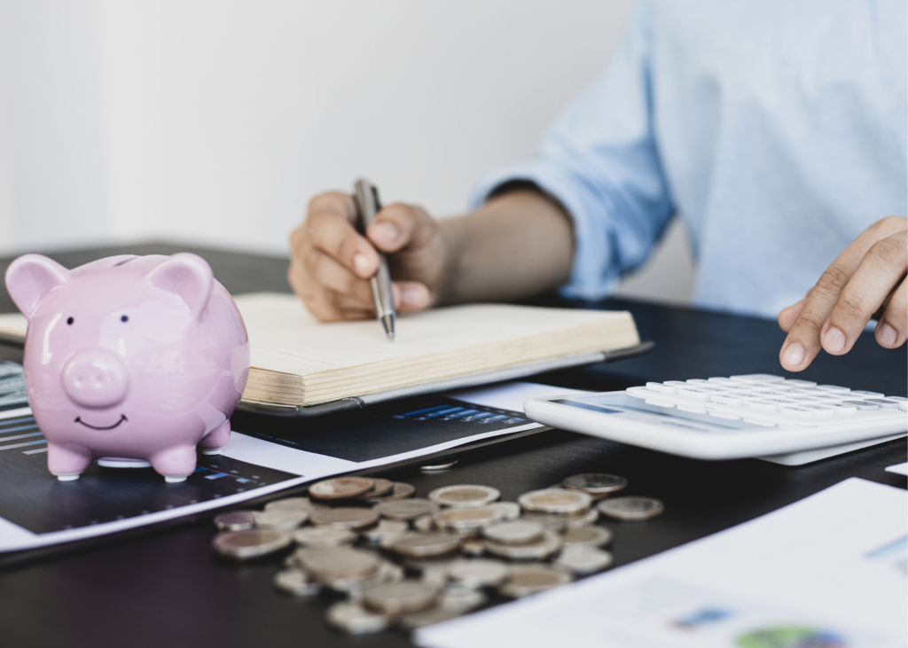 Person sitting at a table, writing in notebook and using calculator. Printouts under notebook, pink piggy bank and coins sitting on table.