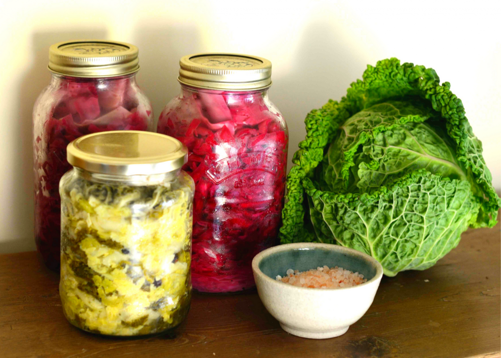 Three mason jars filled with fermented vegetables on wooden table beside whole green cabbage and salt in a bowl.