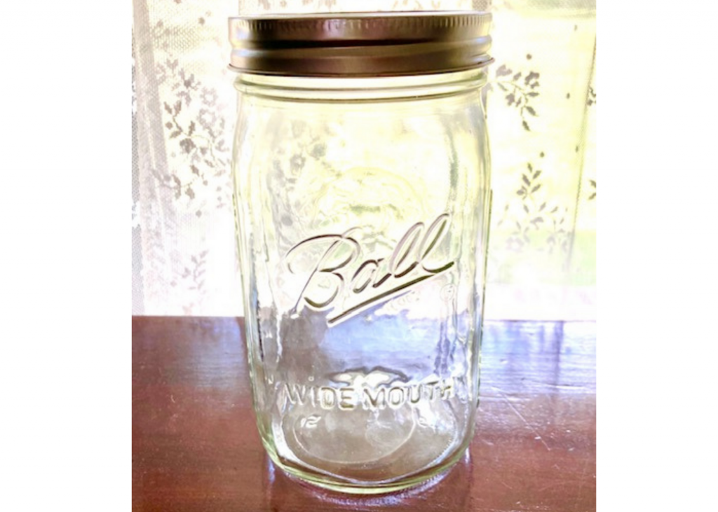 Empty ball wide mouth mason jar with lid on sitting on brown table with lacey curtain background.