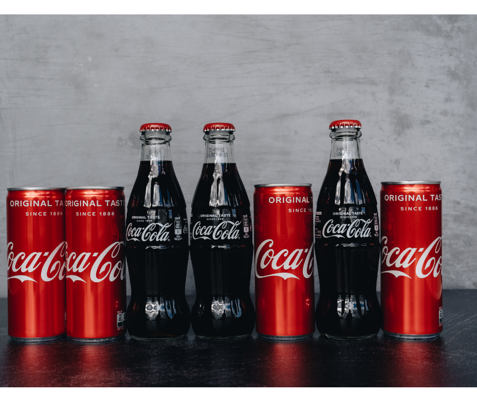 Carbonated Coca-Cola products lined up on black counter with grey backdrop. Two red coca-cola cans, two glass coca-cola bottles, one coca-cola can, one glass coca-cola bottle and one coca-cola can.