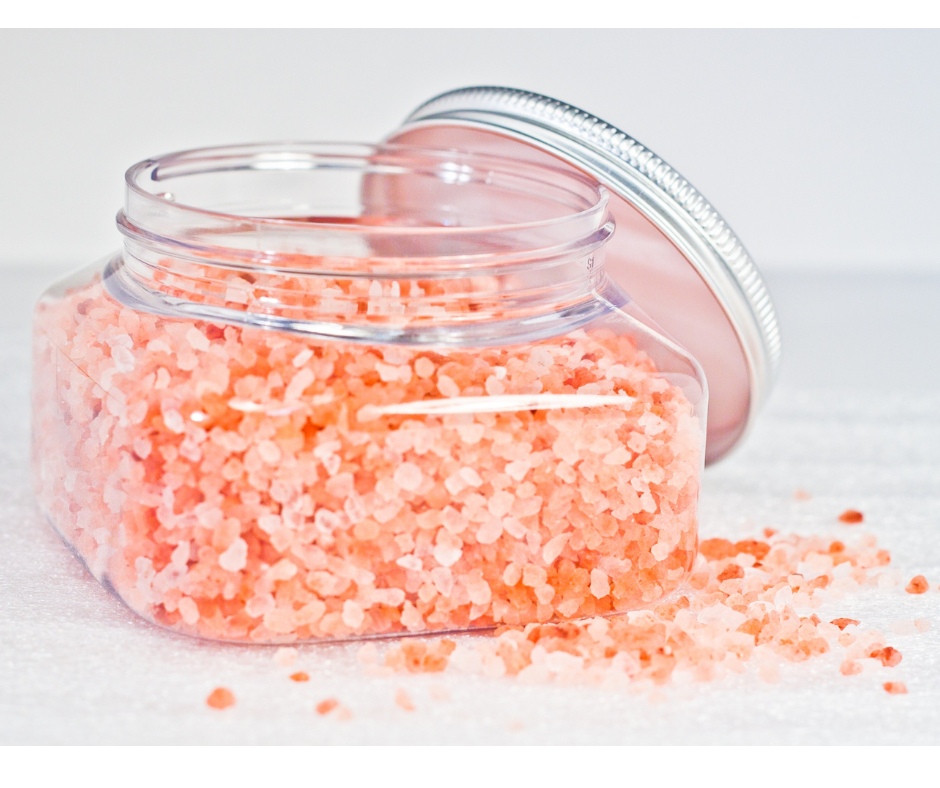 Jar of pink Himalayan salt sitting on a while surface with some salt spilled out and lid leaning against jar.