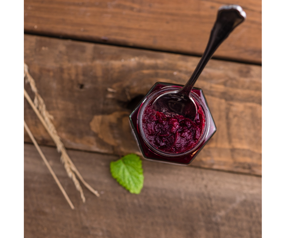 Jar of jam with pectin and spoon in it beside green leaf on a wooden table.
