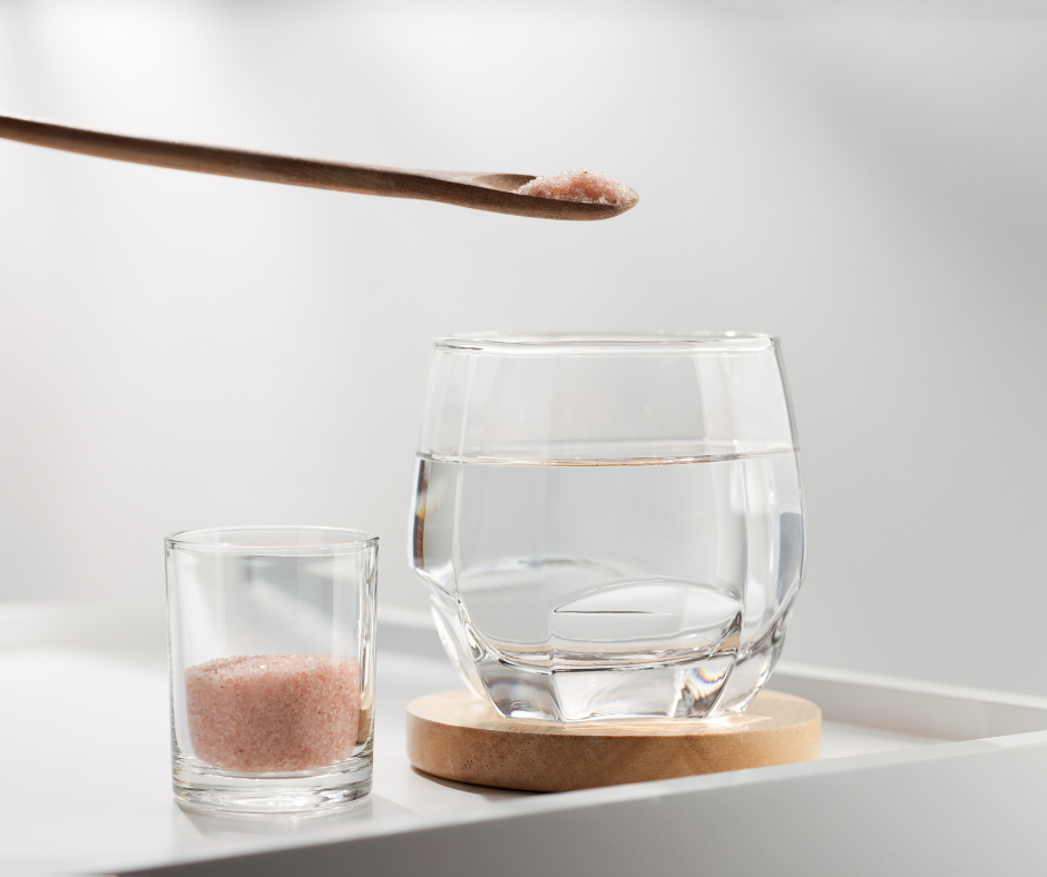 Pink salt in clear glass on white table being measured with a wooden spoon into a glass of water.