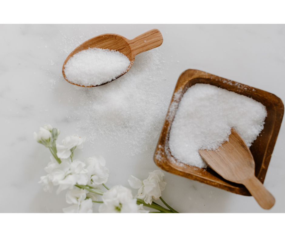 White fermenting salt sitting in wooden square dish with two wooden scoops. Salt spilled on white table from one of the wooden scoops and white flowers placed in the corner.