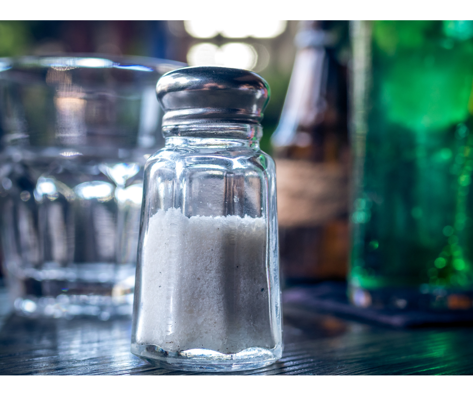 White table salt in salt shaker on wooden table with an empty glass in the rear left and a glass to the right with green liquid in it.