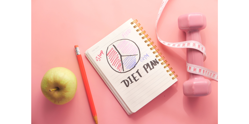 Green apple, pencil, small weight with measuring tape wrapped around it and notebook with diet plan written on it.