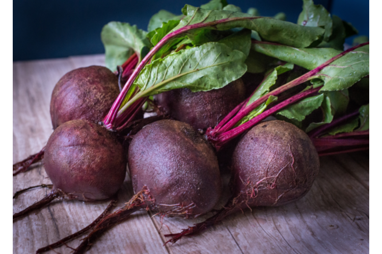 Five purple beet roots with attached green tops laying on a wooden table.