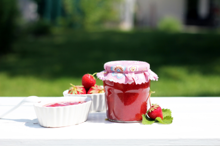 Jar of strawberry jam sitting on a white table outside with strawberries sitting beside it and a white bowl of jam.