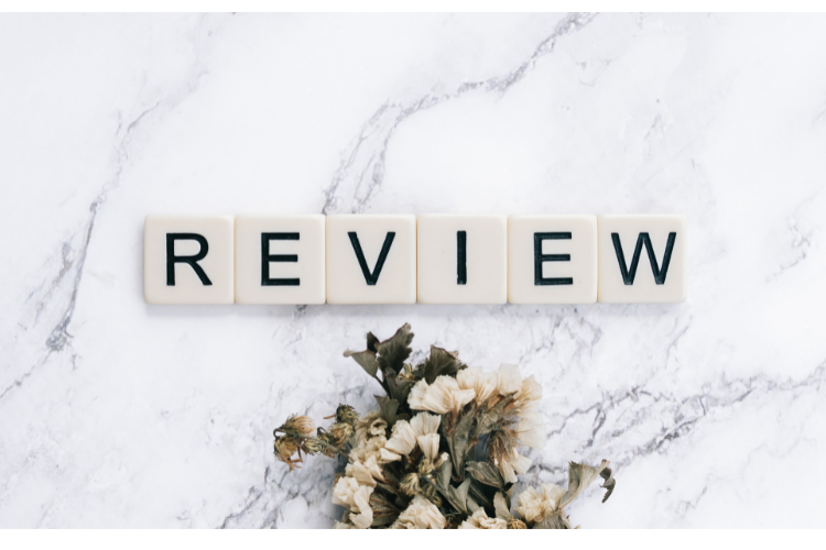 Scrabble letters REVIEW sitting on a white marble table with white wilted flowers underneath.
