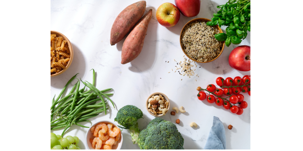 Various vegetables, herbs, grains and meat sitting uncut on a white surface.