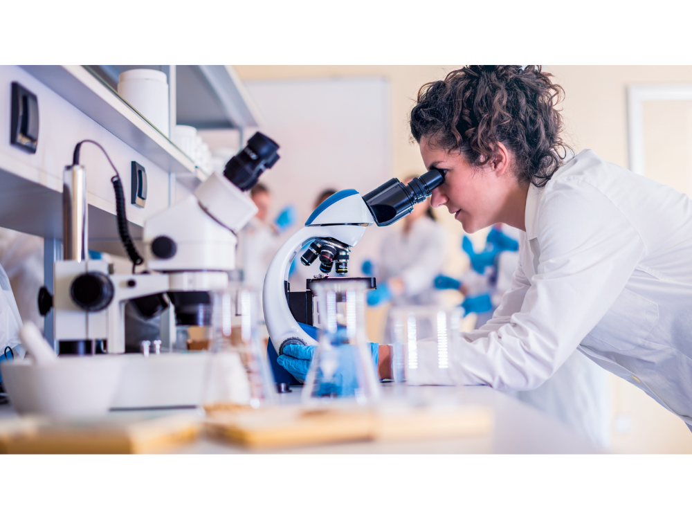 Female researcher looking into microscope with white lab coat on and blue gloves. Other equipment out of focus on the work bench.