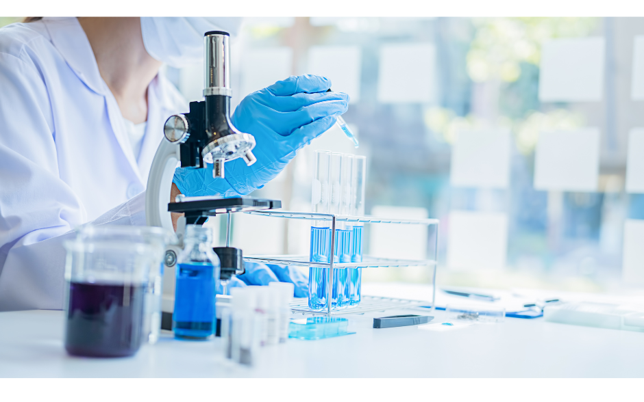 Researcher with mask on putting blue liquid into test tubes that are in container on table with other research equipment.