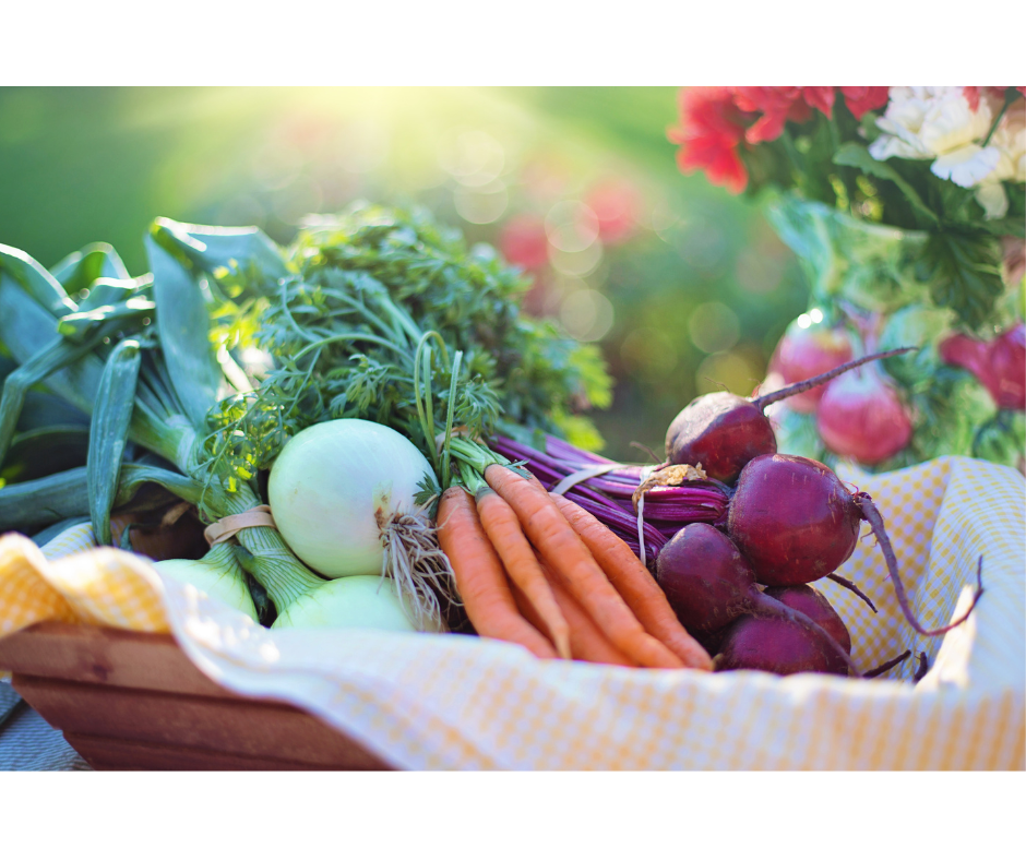 Fresh garden vegetables in wooden container with yellow checkered towel inside, flowers behind and sunlight hitting the scene from behind