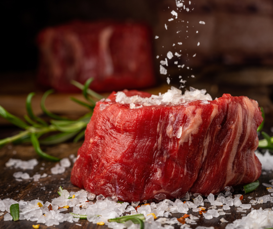Raw chunk of meat with nitrates and salt being added to it on a wooden cutting board with green herbs and meat in the background