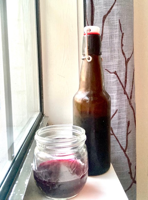 amber swingtop bottle with beet kvass in it and small glass jar in front with fermented beet kvass in it. All sitting on white wood next to a window